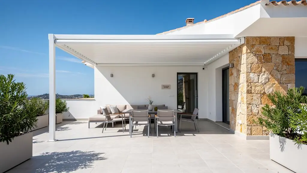 Vista amplia de una terraza mediterránea con toldo logia de estructura blanca integrado en una vivienda contemporánea, mobiliario discreto al fondo y luz natural intensa